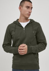 Olivovo zelená mikina na zips s kapucňou a sťahovacou šnúrkou, so žebrovanými manžetami a lemom. Vyrobená z mäkkého materiálu; na boku je malá logová nášivka.