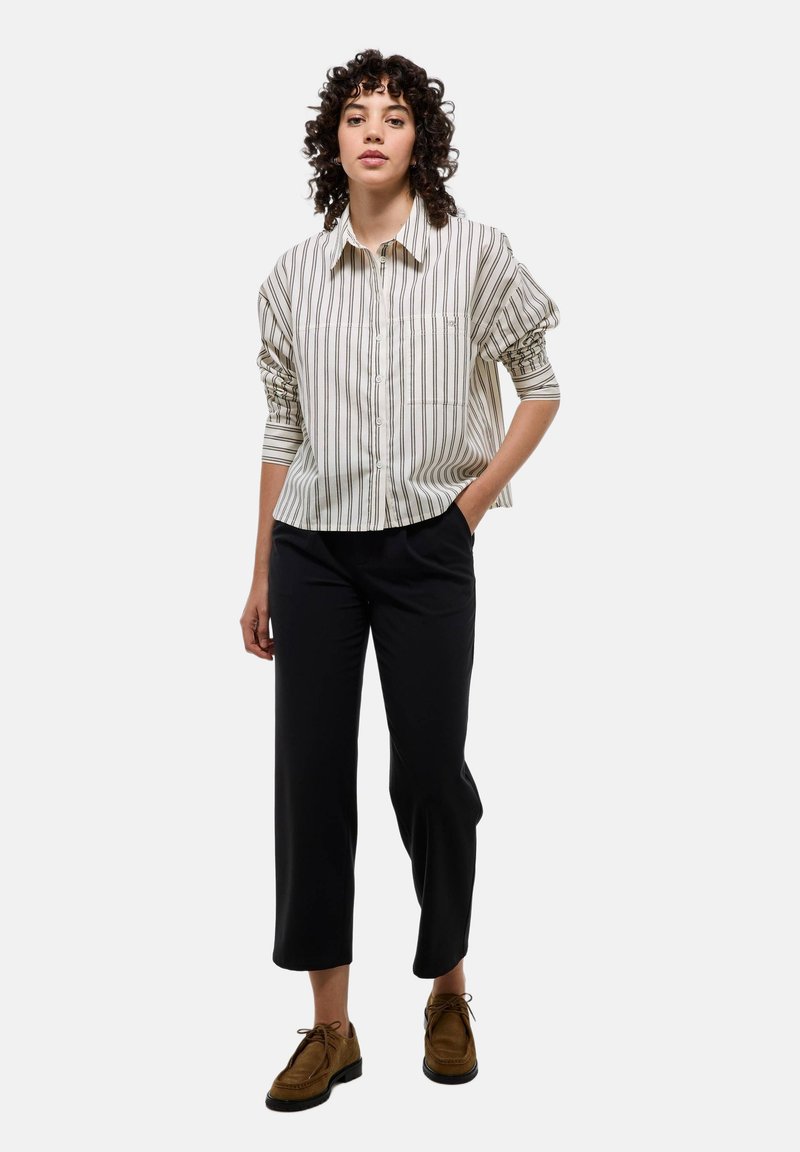 Chemise à rayures beige clair et crème, coupe décontractée avec des manches froncées. Associée à un pantalon noir et des chaussures à lacets marron.