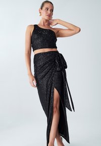 Black two-piece outfit featuring a one-shoulder cropped top and a high-slit wrap skirt, both covered in sequins for added texture.
