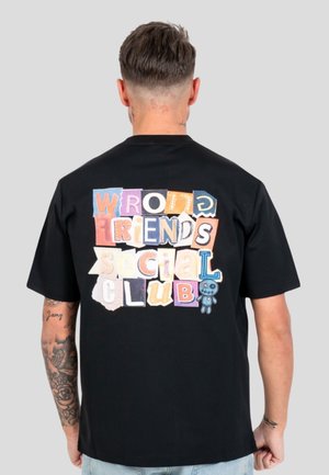 Hombre con pelo corto y tatuajes que lleva una camiseta negra con letras de estilo parche coloridas que dicen "Wrong Friends Social Club" en la espalda.
