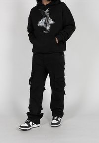 Sudadera negra con gráfico de ángel, bolsillo frontal tipo pouch; pantalones cargo negros con múltiples bolsillos; zapatillas en blanco y negro. Fondo simple.