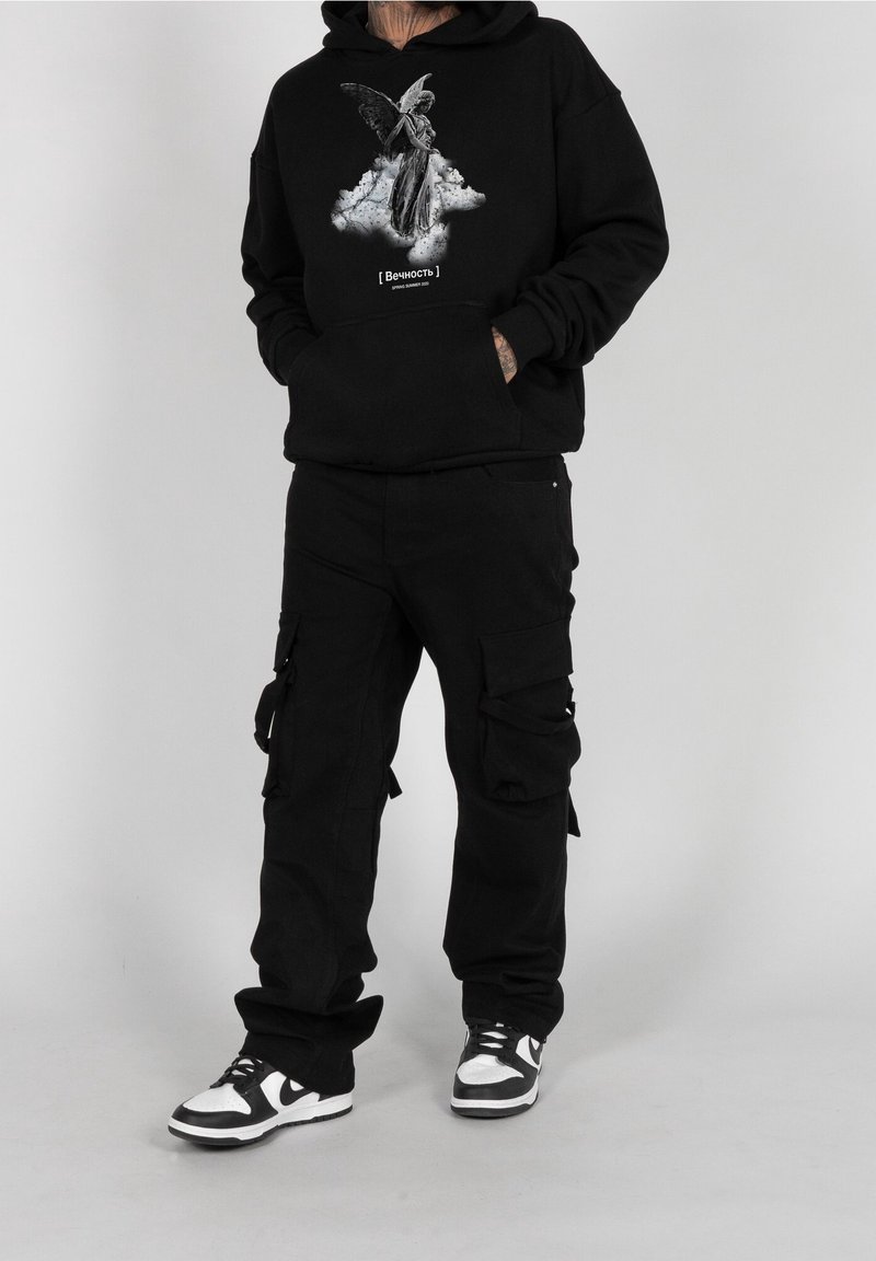 Sudadera negra con gráfico de ángel, bolsillo frontal tipo pouch; pantalones cargo negros con múltiples bolsillos; zapatillas en blanco y negro. Fondo simple.