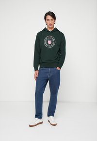 Grønn hettegenser med et sirkulært logo. Sammen med blå denimjeans og hvite sneakers. Enkelt design med en avslappet passform.