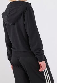 Felpa nera con una vestibilità comoda, caratterizzata da un grande cappuccio, maniche lunghe e polsini a coste. Abbinata a pantaloni neri con strisce laterali bianche.