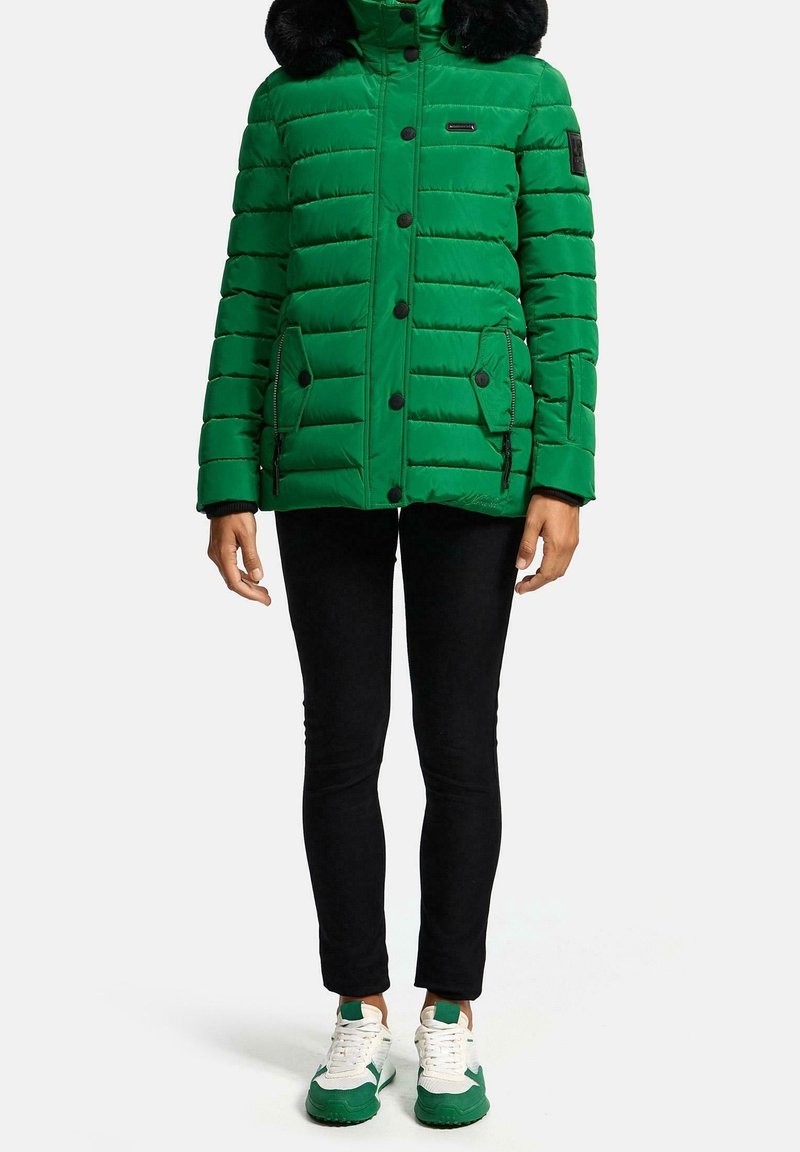 Chaqueta acolchada verde con capucha de piel sintética, bolsillos laterales con cremallera y botones a presión negros. Llevada sobre pantalones negros y zapatillas con detalles en verde.