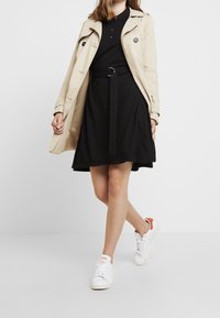 Trench beige superposé sur une robe noire avec une ceinture. Baskets blanches avec des accents orange. Design simple et classique avec des lignes épurées.