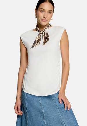 Femme portant un haut blanc sans manches, un foulard léopard autour du cou, des boucles d’oreilles créoles dorées et une jupe en jean bleue, debout devant un fond uni.