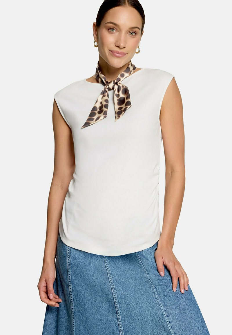 Femme portant un haut blanc sans manches, un foulard léopard autour du cou, des boucles d’oreilles créoles dorées et une jupe en jean bleue, debout devant un fond uni.