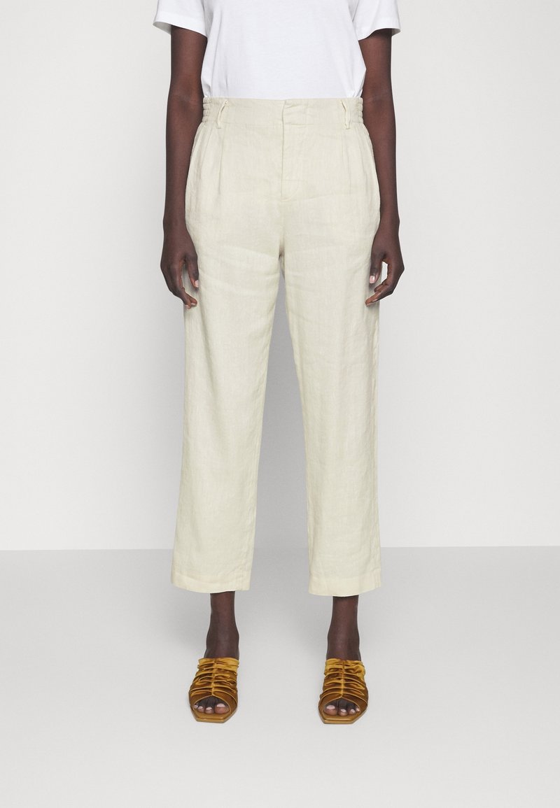 Personne portant un pantalon beige ample taille haute, un haut blanc à manches courtes et des sandales à talons ouvertes couleur moutarde, debout devant un fond uni.