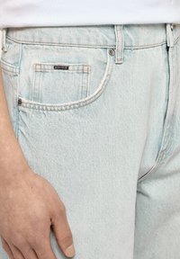 Jeans in denim azzurro chiaro con una texture leggermente sbiadita, dotati di una tasca frontale e di una piccola etichetta nera cucita sulla vita.