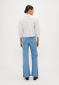 Bluză albă cu mâneci bufante, împodobită cu broderie florală albastră, asortată cu blugi evazați de culoare albastru deschis. Blugii au fante laterale și un croi clasic.