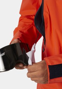 Chaqueta impermeable naranja con detalles en cremallera negra; persona sosteniendo gafas de esquí negras, limpiando la lente con un paño blanco.