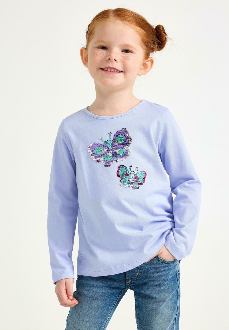 Lyse lilla langærmet skjorte med sommerfugle-sequindesigns i grønblå, lilla og rød. Kombineret med blå jeans. Glat stoftekstur.