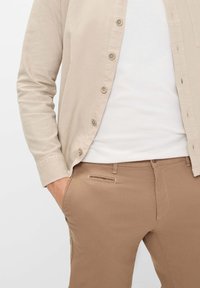 Beige blouse met lange mouwen over een witte T-shirt, gecombineerd met lichtbruine, slim-fit broek. Opvallende details zijn de knoopaccenten.
