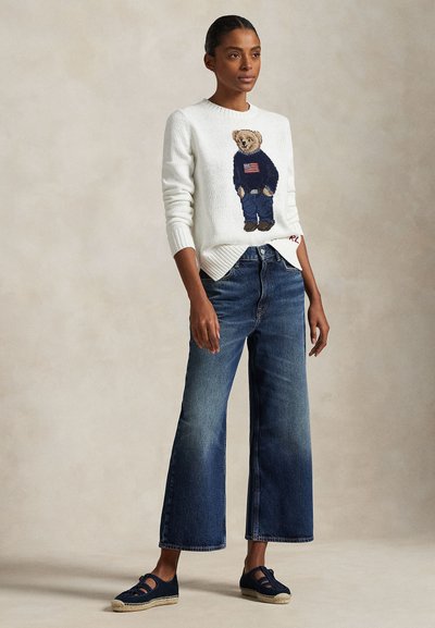 Ženska oblečena v belem puloverju z medvedkom v puloverju z ameriško zastavo, širokih modrih kavbojkah in mornarsko modrih espadrilah, stoji z sproščenimi rokami.