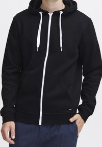 Felpa con cappuccio nera con cerniera, coulisse bianche, tasche frontali e polsini a coste. Realizzata in tessuto morbido, presenta un design pulito e un piccolo logo.