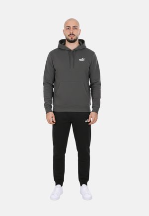 Hombre de pie de frente vistiendo una sudadera con capucha Puma gris oscuro, pantalones deportivos Puma negros y zapatillas blancas sobre un fondo blanco.