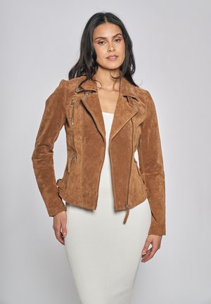 Vrouw draagt een bruine suède bikerjack over een witte, geribbelde, nauwsluitende jurk, staand tegen een effen lichtgrijze achtergrond.