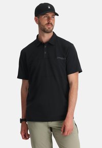 Polo nero con un design a trama, colletto a tre bottoni e un sottile logo "SPYDER" sul lato sinistro del petto. Indossato con pantaloni di colore chiaro.