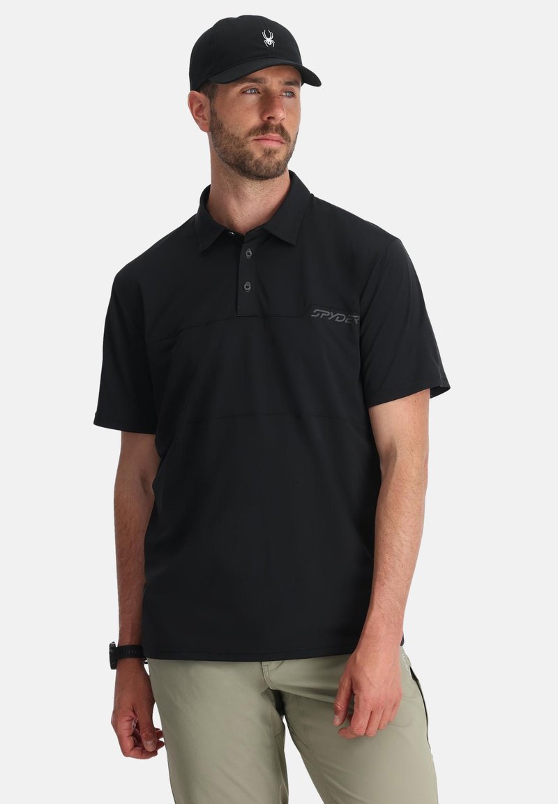 Polo nero con un design a trama, colletto a tre bottoni e un sottile logo "SPYDER" sul lato sinistro del petto. Indossato con pantaloni di colore chiaro.