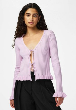 Žena s dlhými kučeravými vlasmi, oblečená v svetlofialovom rebrovanom kardigáne zviazanom vpredu a čiernych širokých nohaviciach, stojaca pred bielym pozadím.