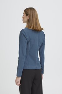 Õlgadeni helepruuni juustega naine seisab küljepealt, kandes keha järgivat sinist pika varrukaga jopet ja musti püksid valge tausta ees.