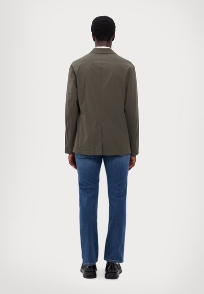 Olive green, lightweight jacket with a single vent at the back, paired with blue jeans and black shoes, viewed from the back.