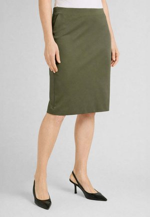 Donna che indossa una gonna a tubino verde oliva al ginocchio e décolleté nere con cinturino sul tallone, in piedi davanti a uno sfondo chiaro uniforme.