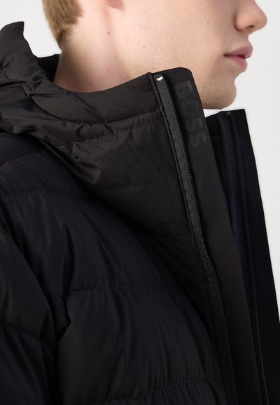 Veste noire rembourrée avec un col haut, dotée d'une fermeture éclair et d'une marque discrète sur le côté. Tissu lisse avec une finition mate.