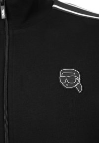 Chaqueta negra con cremallera y textura acanalada, con rayas blancas en los hombros y un parche de logo prominente en blanco y negro.