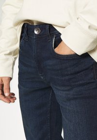 Mörkblå denimjeans med en snäv design, som har en metallknäppning och sidofickor. Omfattande stygn detaljer längs sömmarna.