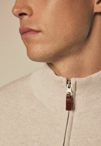 Beige zip-sweater med ribbet krave, med en sølvfarvet lynlås og en brun lædertræk; blød tekstur med en tætsiddende design.