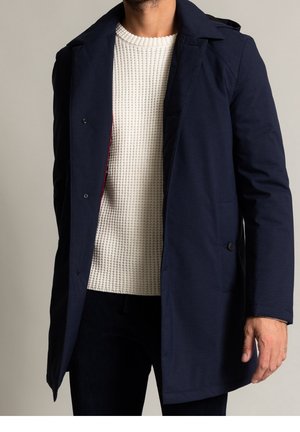 Homme portant un manteau bleu marine sur un pull texturé blanc, debout avec une main détendue le long du corps sur un fond neutre.