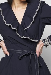 Marinblå omwrap-klänning i lätt bomull med en volangkrage, vita broderidetaljer och ett knytband i midjan. Långa ärmar.