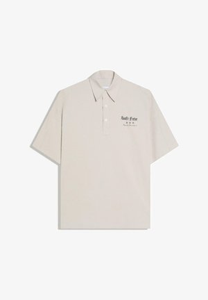 Polo beige à manches courtes avec col et boutons, avec texte noir et petits symboles sur la zone gauche de la poitrine.