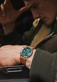 Montre analogique avec un cadran vert, un boîtier en argent et un bracelet en cuir marron. Comprend des sous-cadrans chronographes et un bracelet texturé.