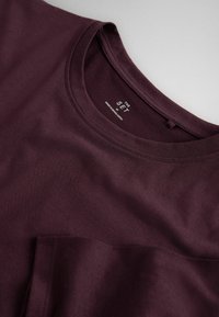 Dark purple crew neck T-shirt showing neckline and part of a front chest pocket, with a visible brand label inside collar.