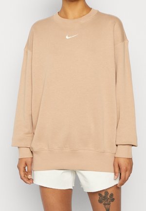 Person iført oversize beige Nike-sweatshirt og hvide shorts, der viser en sort blomster-tatovering på venstre lår.