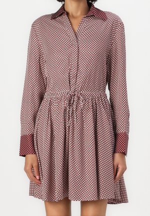Vrouw die een lange mouwen, knielange jurk draagt met een bordeauxrode en witte geometrische print en een strik in de taille, staand tegen een effen achtergrond.
