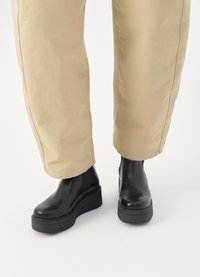 Boots negre cu platformă la glezne, cu un design elegant, realizate din material lucios, având panouri laterale elastice și o talpă groasă neagră, asortate cu pantaloni bej.
