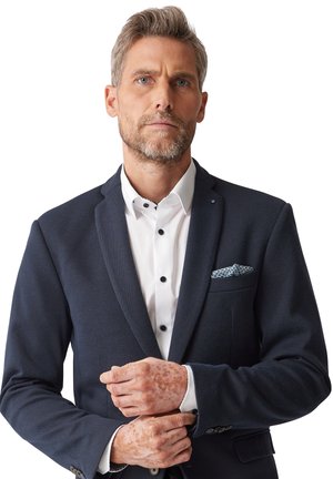 Marineblazer aus strukturiertem Stoff, mit Reverskragen, einreihigem Knopfverschluss, weißem Hemd, schwarzen Knöpfen, gemustertem Einstecktuch und sichtbaren Hautdetails.