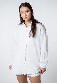 White button-up shirt made from textured fabric, featuring long sleeves, a collar, and pearl-like buttons. Worn over light shorts.