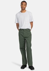 T-shirt gris à manches courtes avec une poche avant, associé à un pantalon droit vert et des chaussures noires. Design simple, couleurs unies.