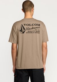 T-shirt en coton beige avec un col rond, featuring une impression graphique noire au dos, comprenant un logo en diamant et du texte.