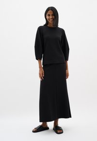 Sort pullover med faldende ærmer, parret med en matchende lang nederdel og flade sorte sandaler; glat tekstur og enkel silhuet.