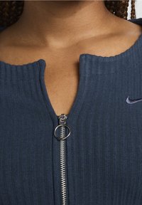 Marineblaues geripptes Oberteil mit einem Reißverschluss und einem runden Ring-Zipper, mit einem runden Ausschnitt und dezentem Nike-Logo auf der linken Seite.
