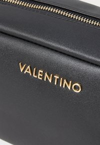 Melnas ādas soma ar zelta tonī ievelkamu rāvējslēdzēju un uzspiestu "VALENTINO" logo priekšpusē, ar gludu tekstūru un minimālistisku dizainu.