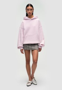 Felpa oversize rosa chiaro con tasca frontale e cordino. Abbinata a una corta gonna fantasia e scarpe con tacco bianco.