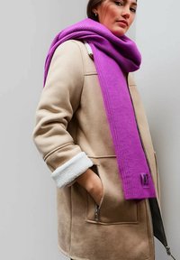 Cappotto in suede beige con bordo in shearling bianco e tasche frontali, abbinato a una vivace sciarpa di lana viola.