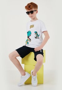 Garçon aux cheveux roux portant des lunettes de soleil noires, un t-shirt blanc avec un graphisme de dinosaure, un short noir et des baskets blanches, assis sur des cubes jaunes empilés.